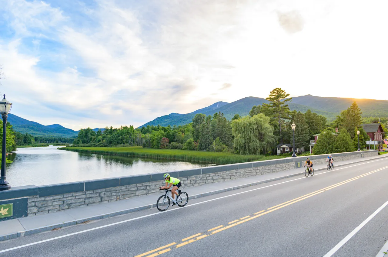 A trio of cyclists cross a stone bridge over a calm river without mountains in the background.