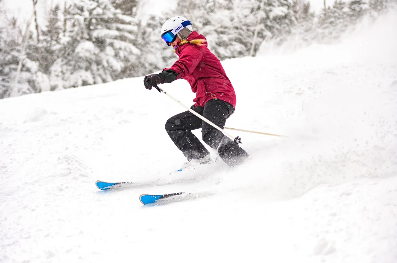 A skier in black and red gear flies down a snowy mountain with snow-covered trees in the background.