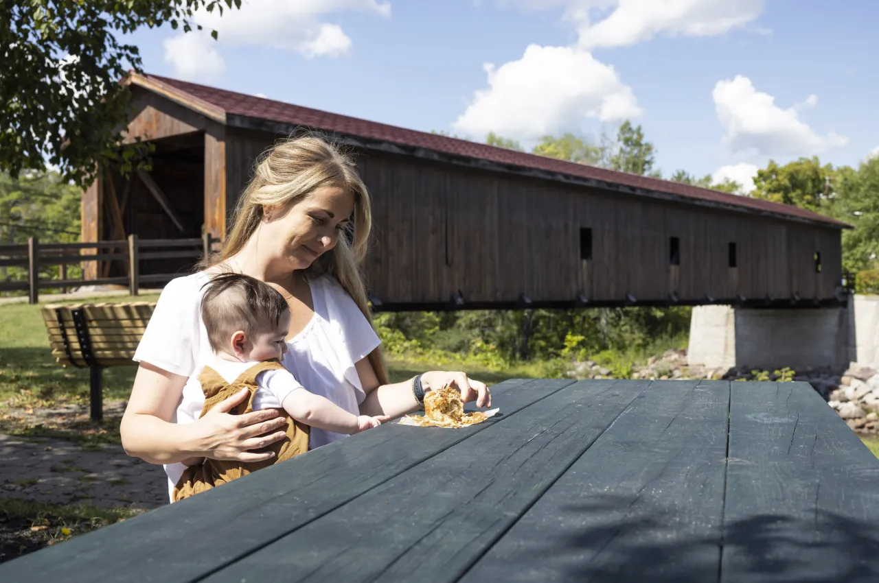 A woman with her baby at a picnic table next to a covered bridge