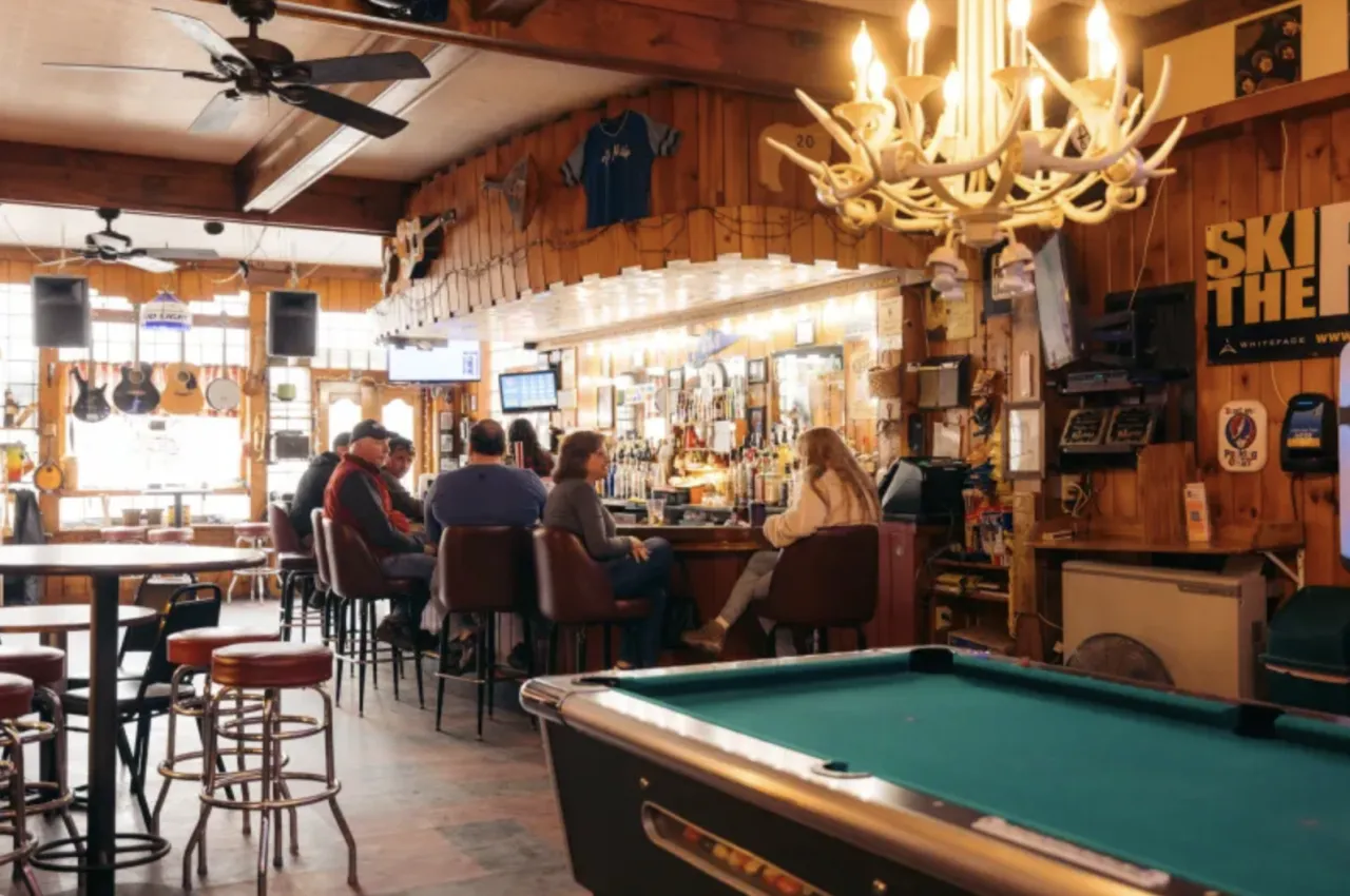The interior of a rustic bar with antler chandelier over the pool table, adults sitting at the bar, and guitars hanging in the bright window.