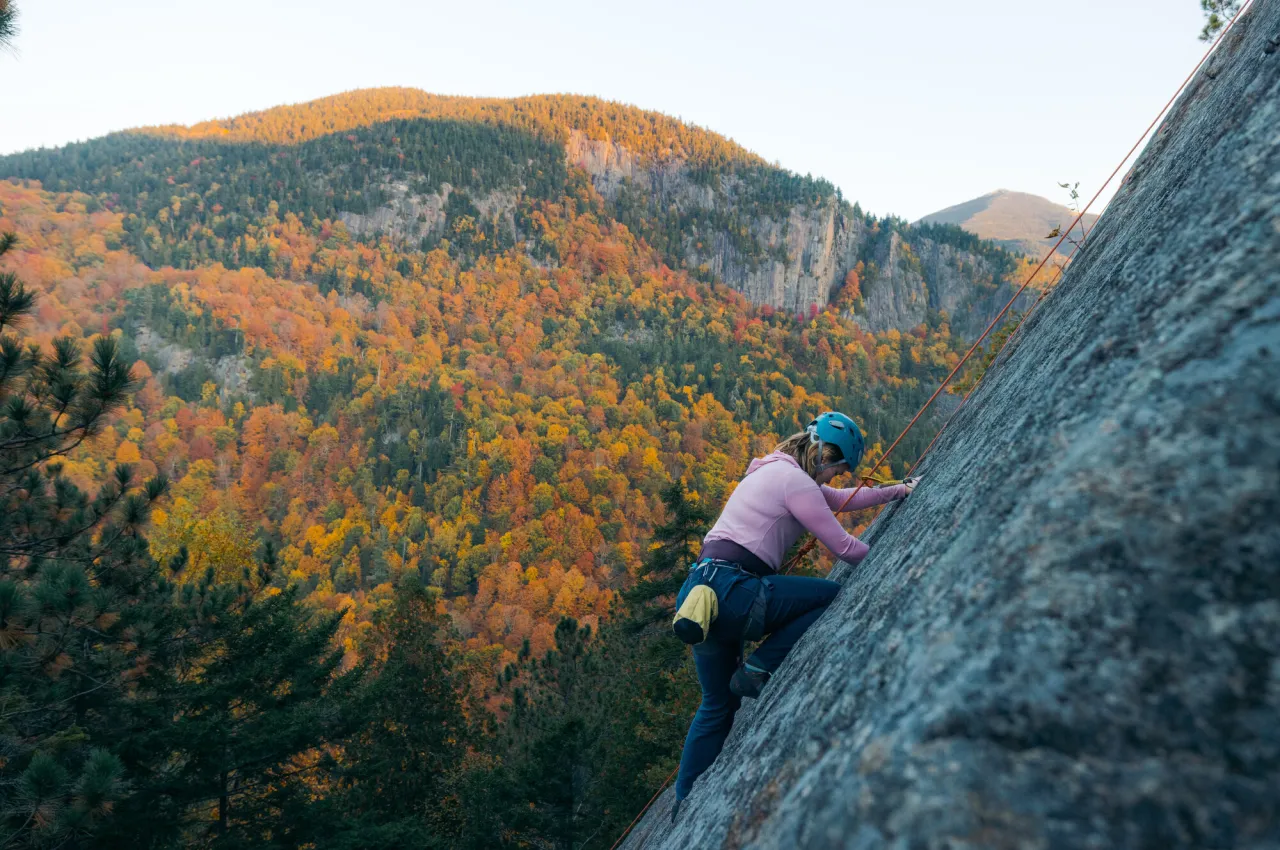 Woman in pink rock climbs slab with fall foliage covered mountain in background