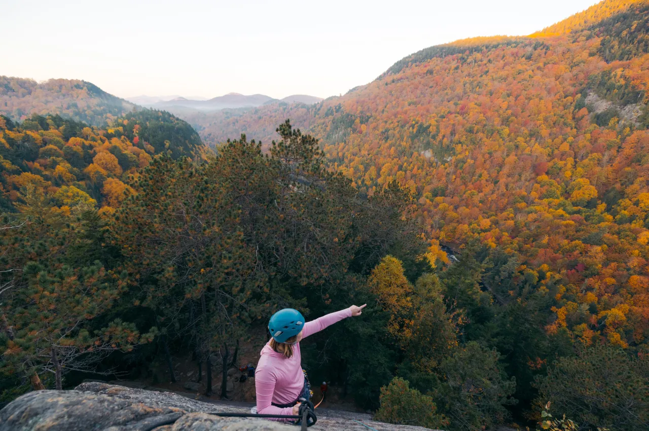 Rock climber at top of a climb points to view of fall foliage covered mountains