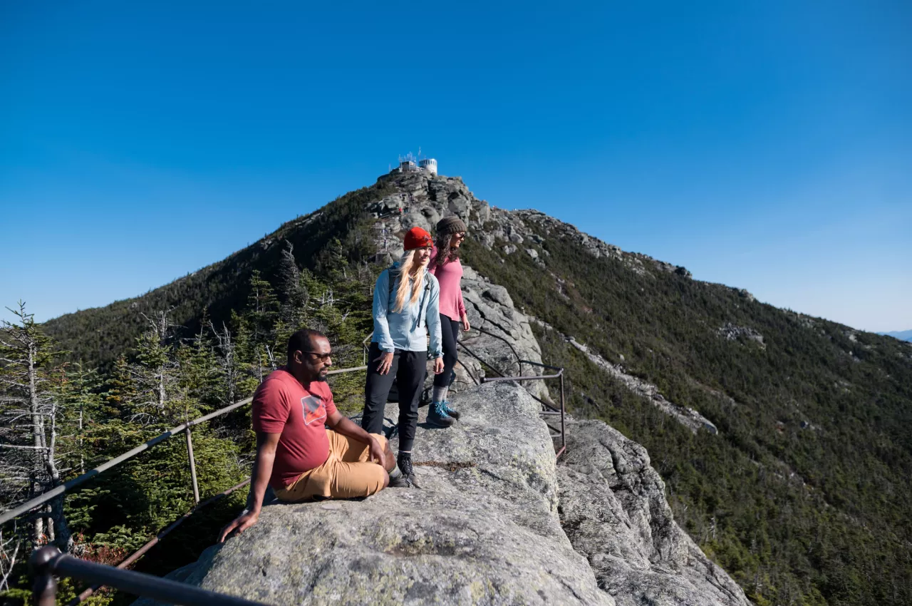 Three adults stand on a rocky ridge below a mountain summit.