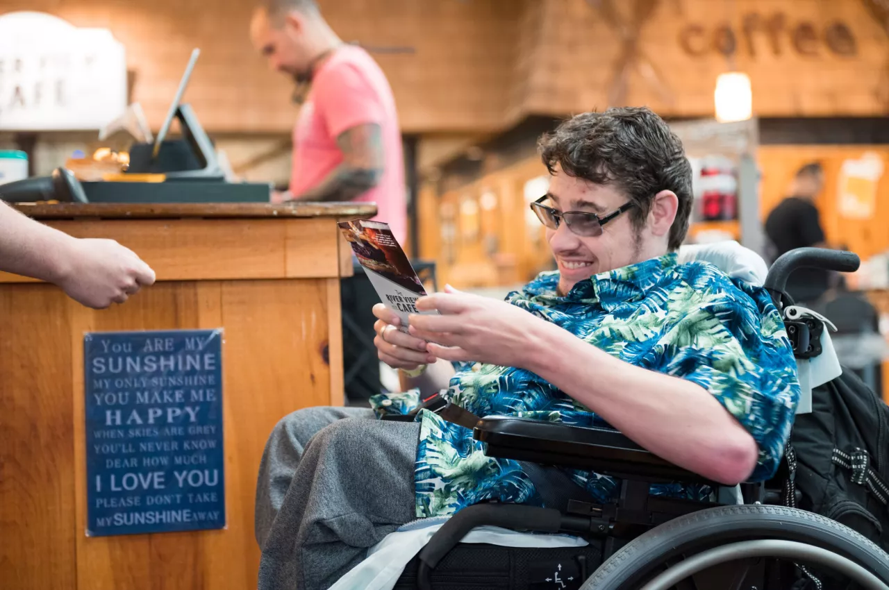 A young man in a motorized wheelchair smiles and reads a brochure next to a ticket desk.