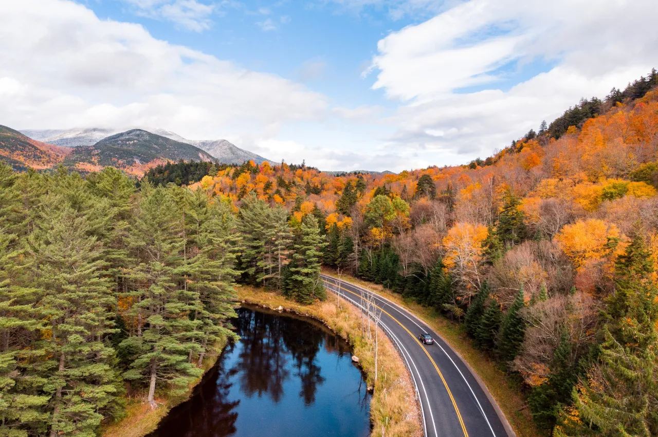 A road runs alongside a river with fall foliage forest on either side and mountains in the distance