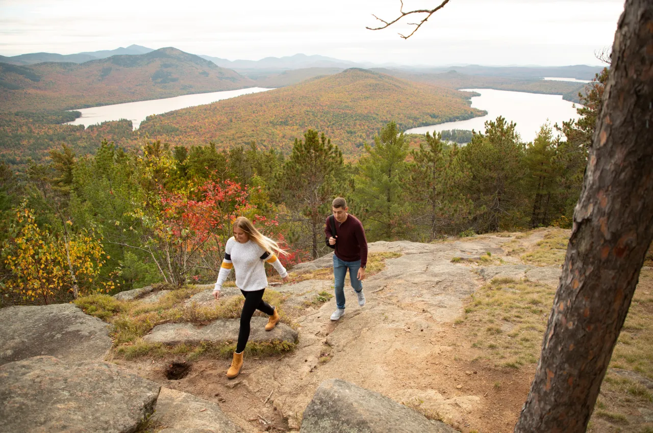 A man and woman walk along a rock outcrop with a fall view of mountains and lakes in the background