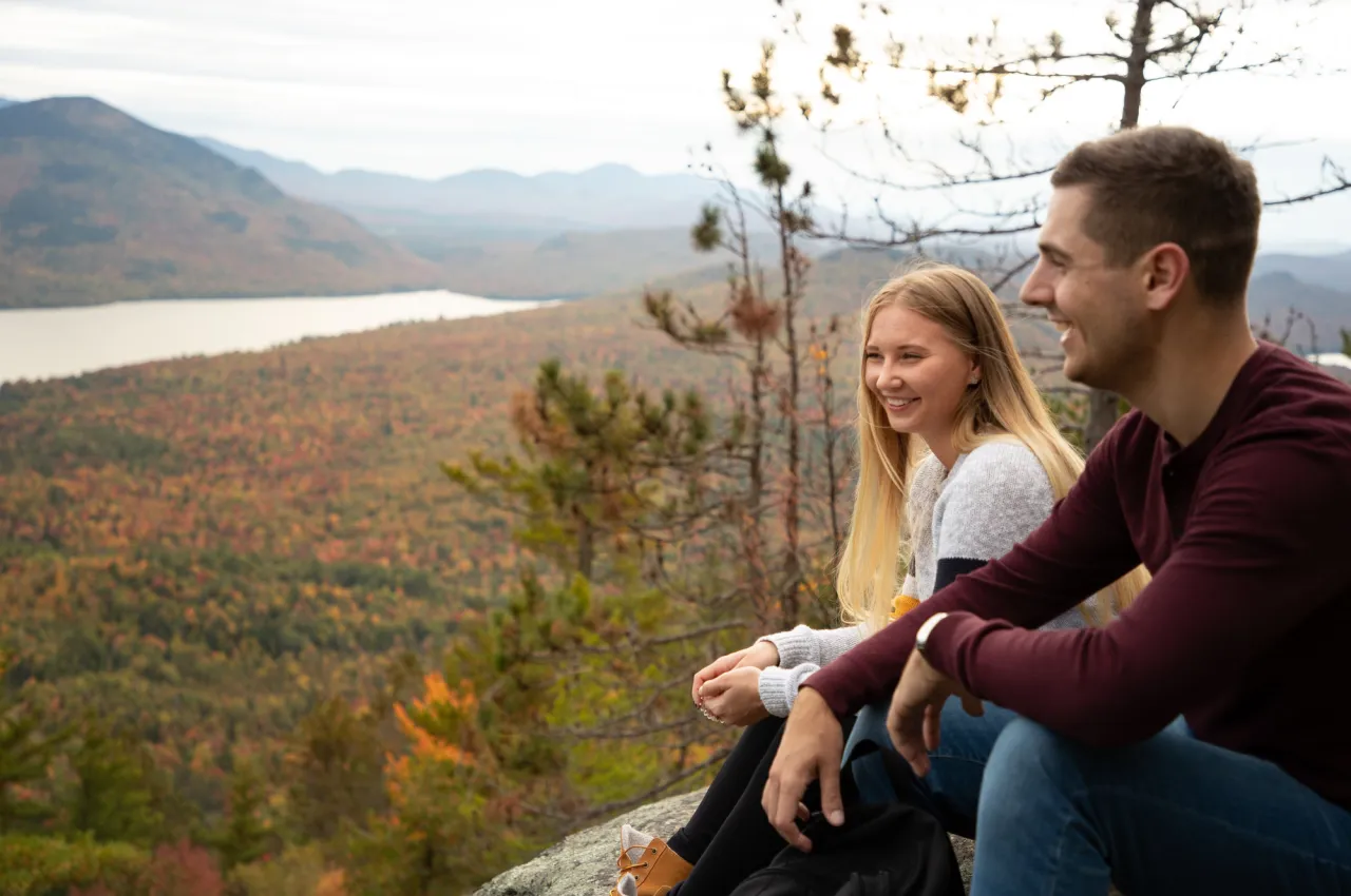 A man and woman sit atop a mountain looking at fall view of mountains and lakes