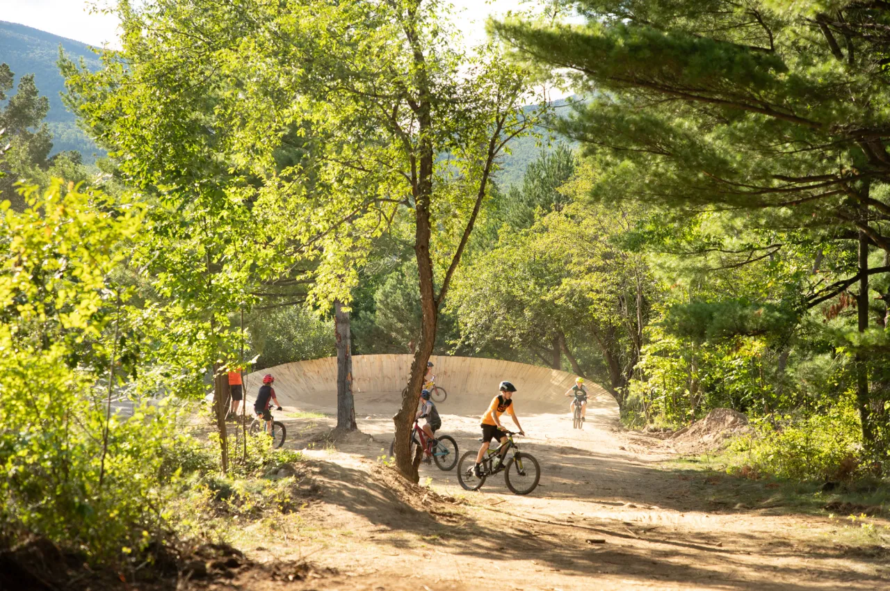 Several young bikers ride around bike park with features in background on sunny day