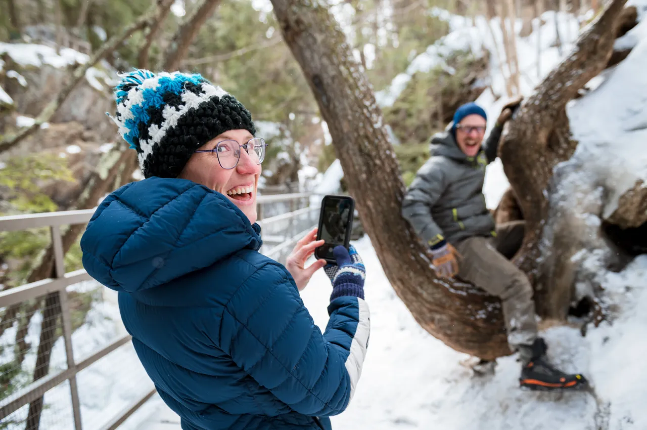 Woman dressed for winter holds phone while looking back over her shoulder while man sits on tree smiling