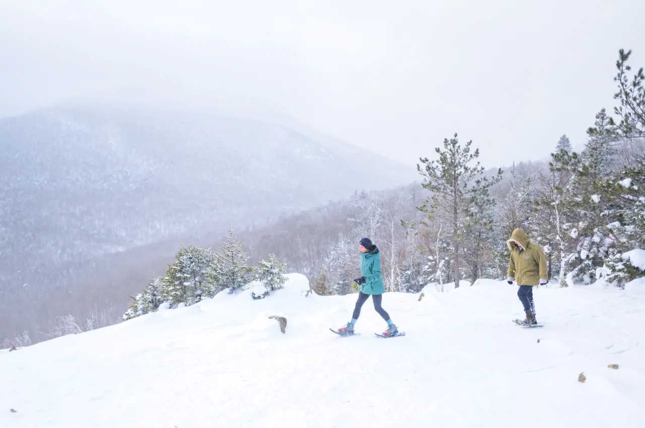 Two people walk along a rock outcrop with a winter view of faint mountains through snow.