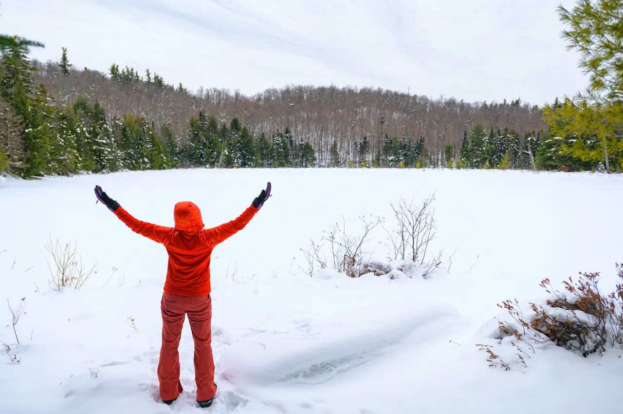 Person in red snow suit stands facing away with arms stretched overhead looking at snowy pond