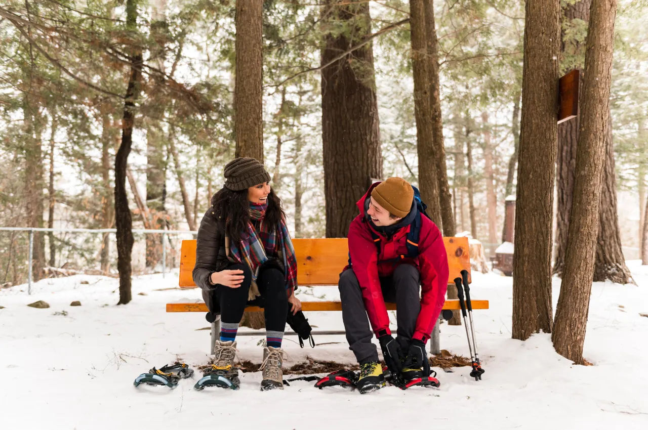 Man and woman sit on bench in snowy woods putting on snowshoes