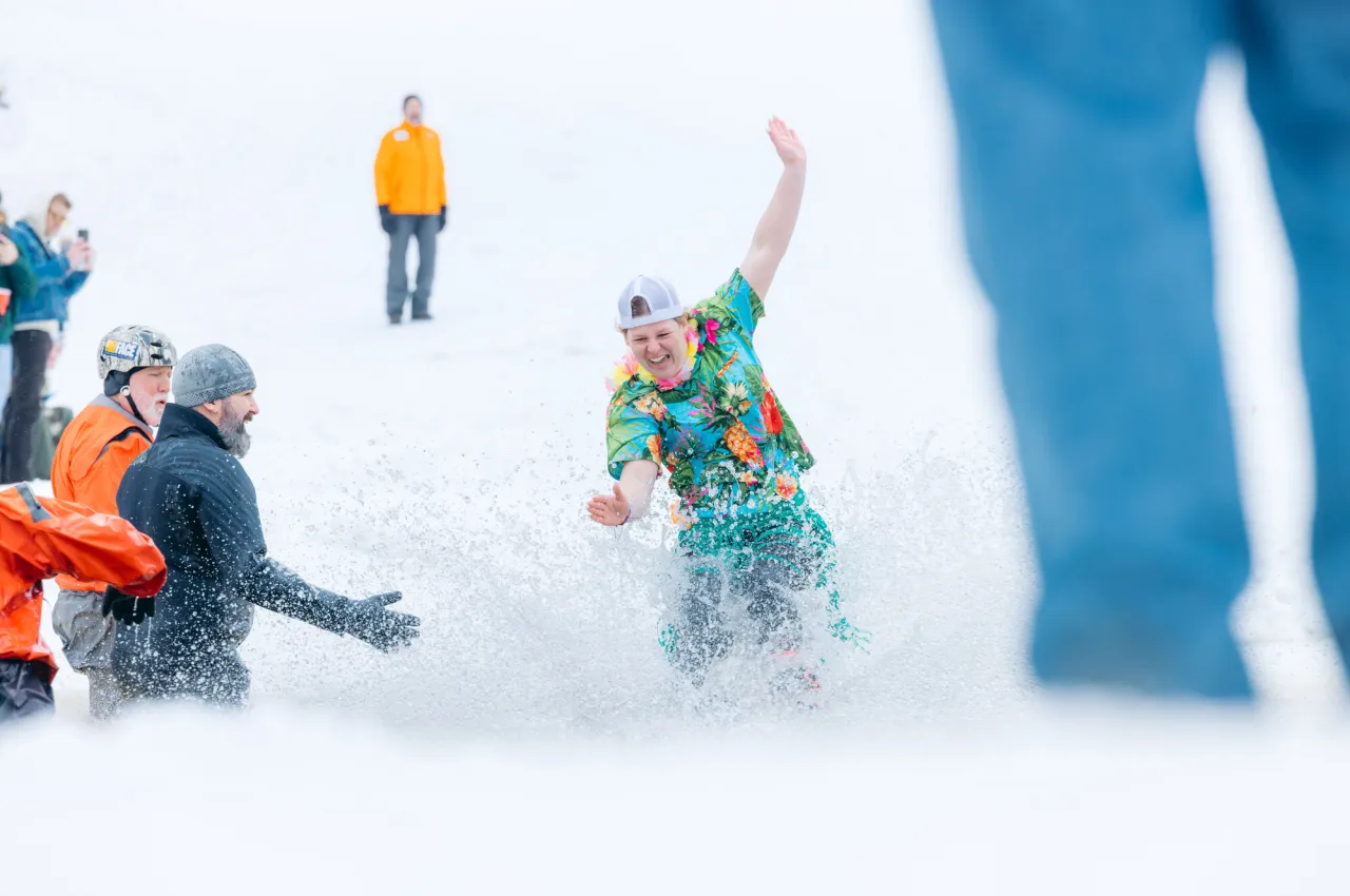 Man in short sleeve Hawaiian shirt skis over pond surrounded by snow