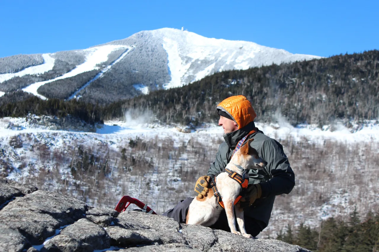 Person holds onto dog on summit with snowy Whiteface Mountain in background