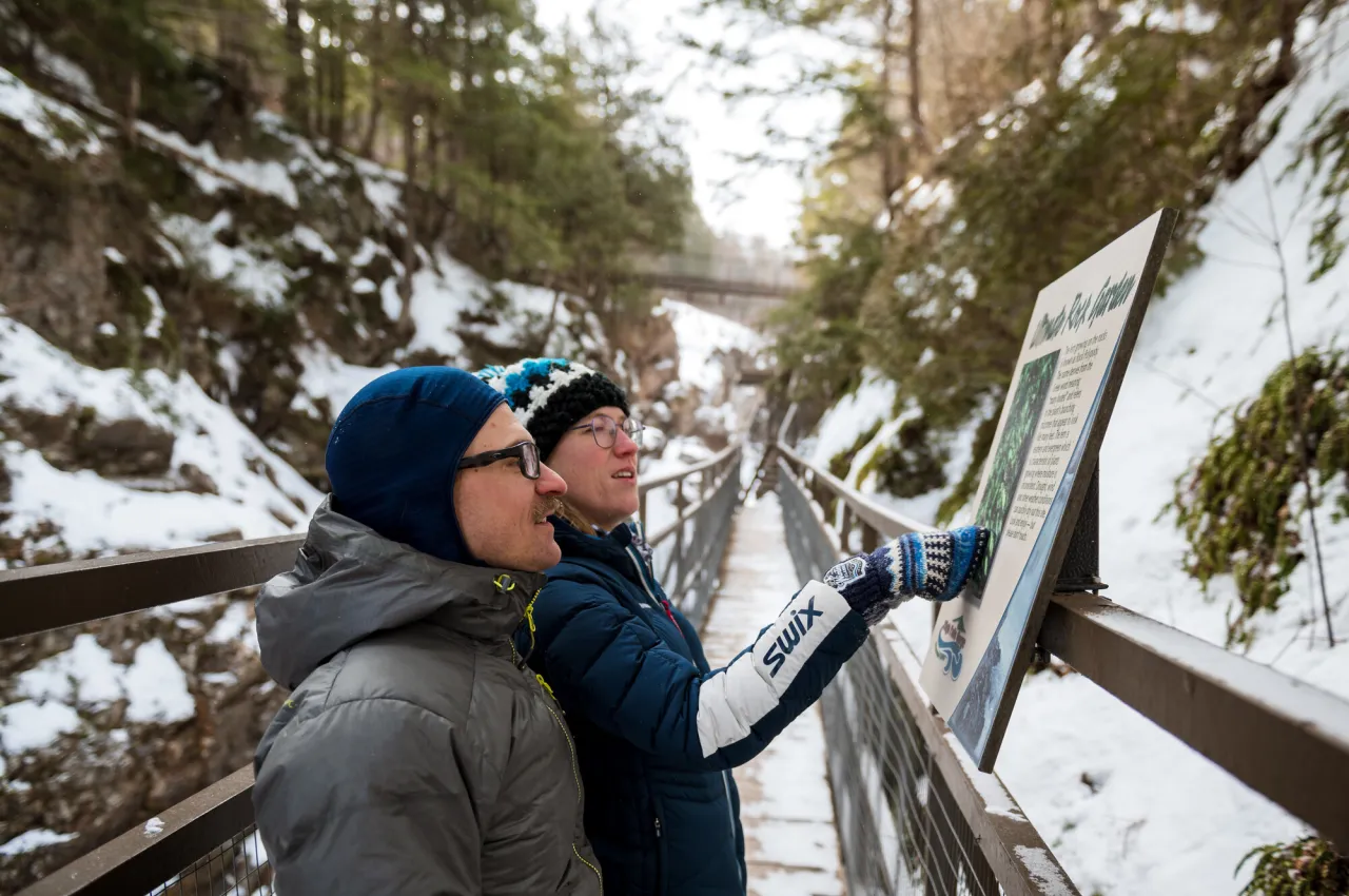 Man and woman in winter clothing look at informational sign along boardwalk