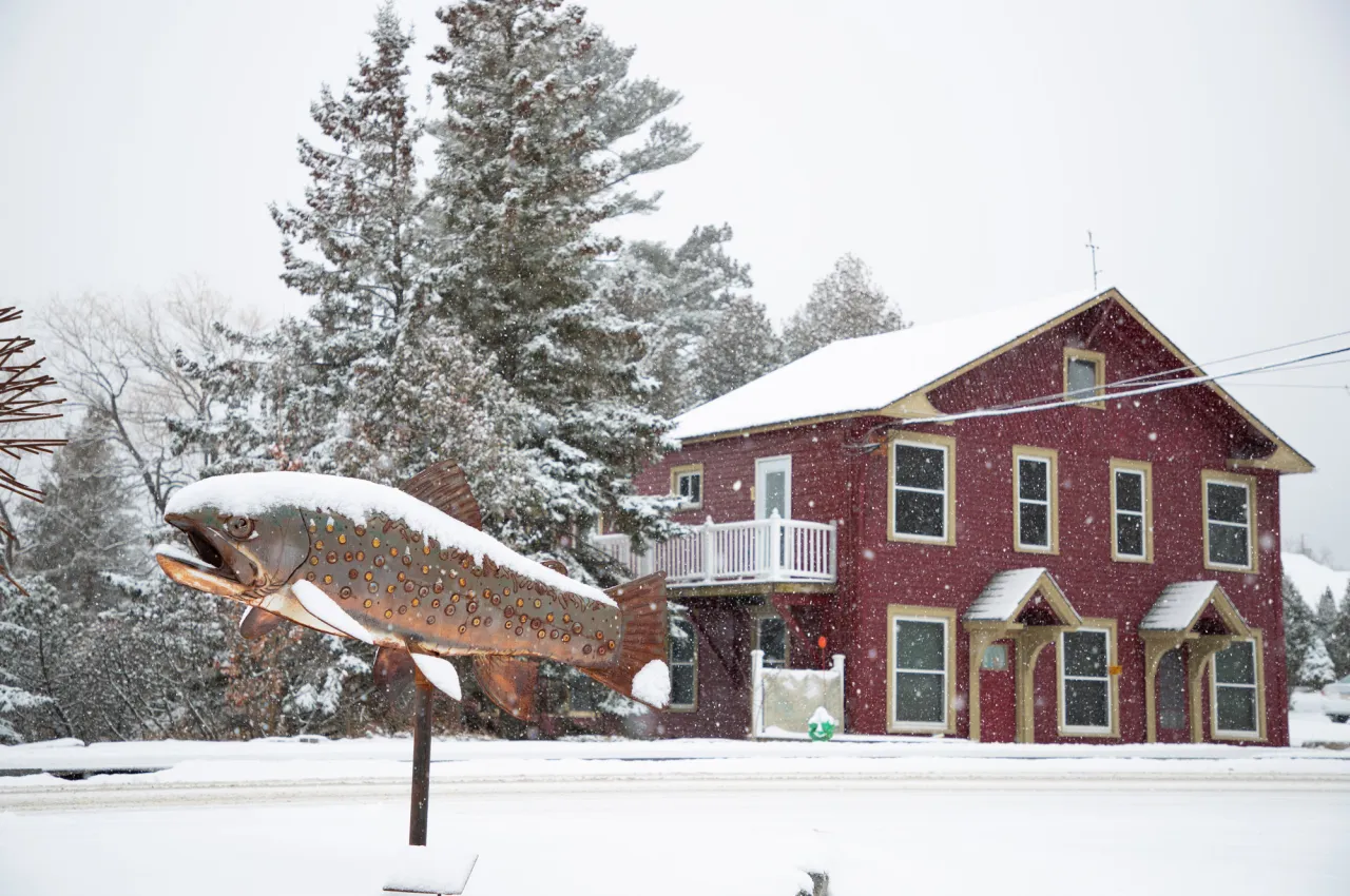 A metal fish sculpture covered in snow in front of a red house