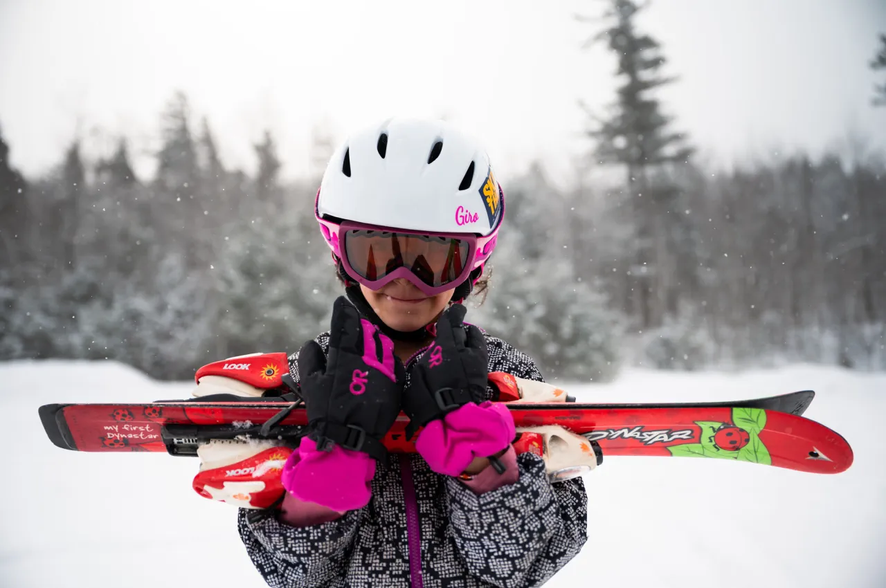 Little girl in ski helmet, goggles, and gear holds skis