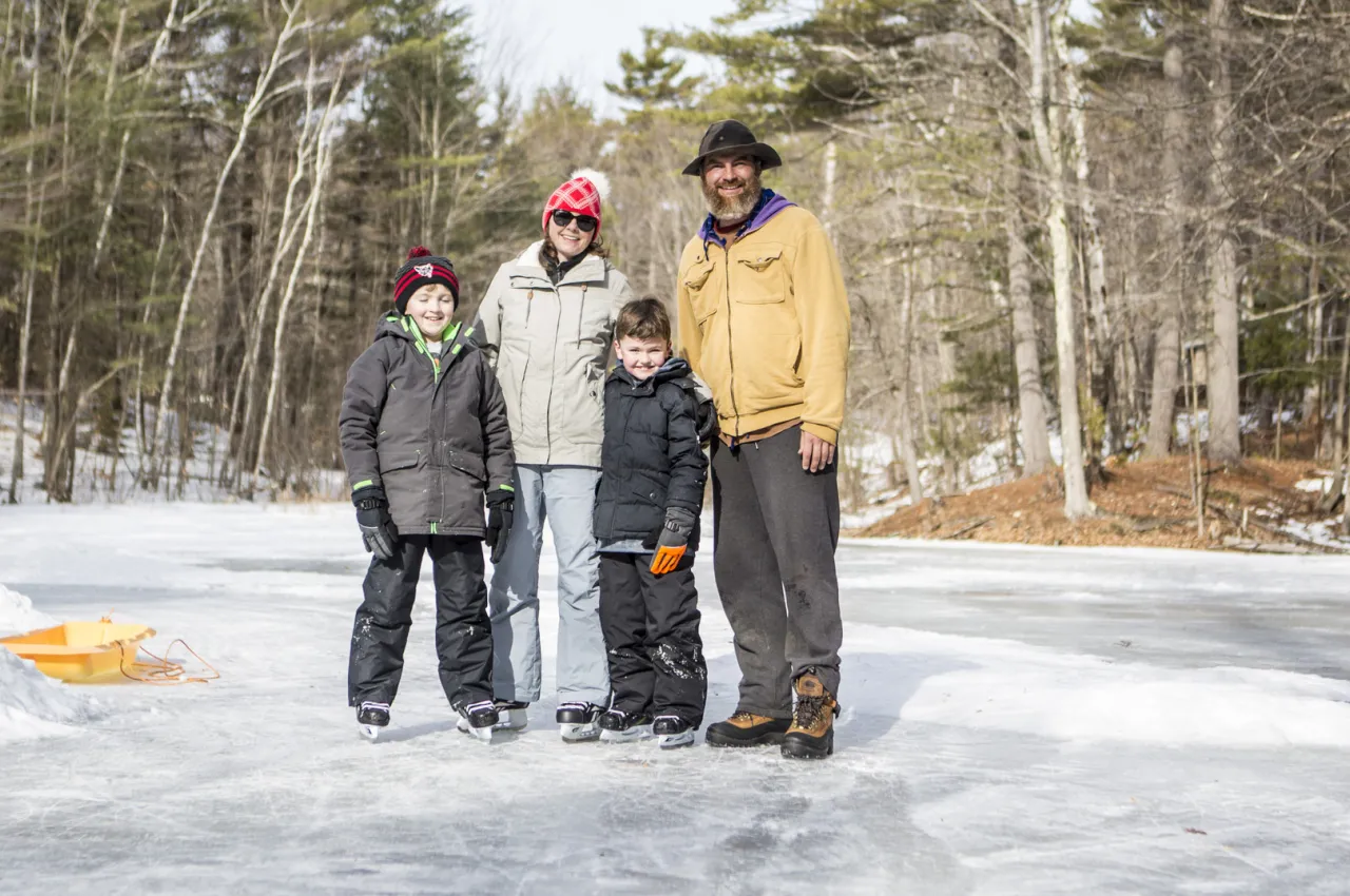 Family of four dressed in winter gear with three people in ice skates stand on ice in forest