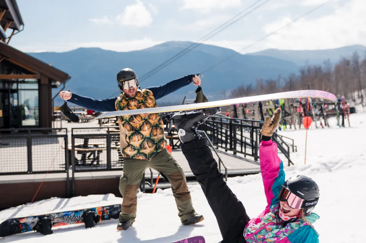 Skier lays on ground with one ski in air while man in pineapple costume raises arms behind