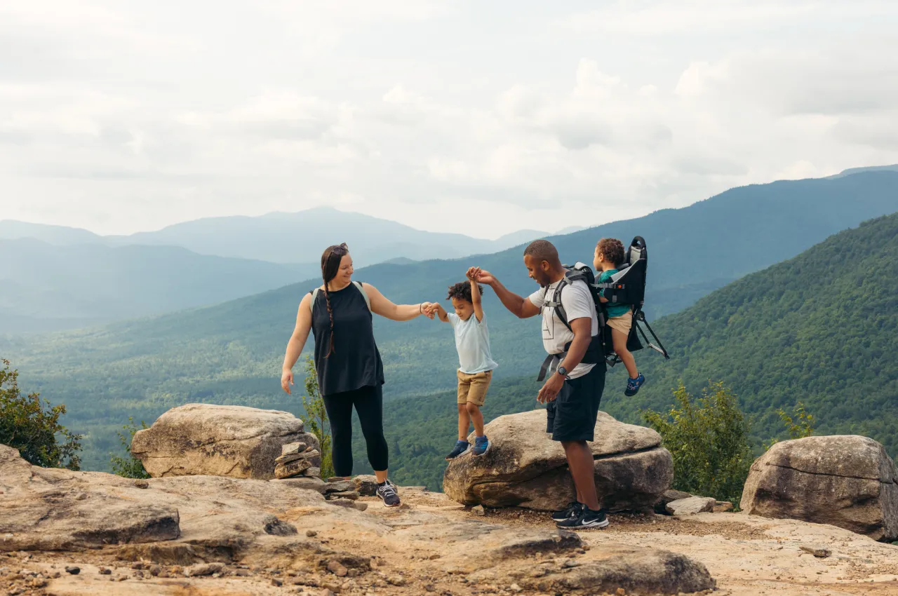 A mother and father wearing a baby in a hiking pack swing toddler off rock on mountain summit