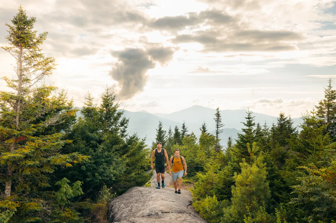 Two men walk on rocky path between trees with mountains in background