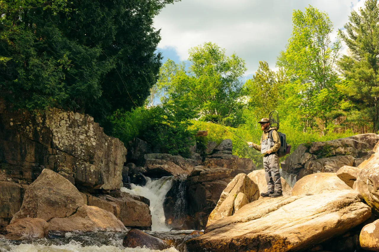 A man in wades stands on river rock fly fishing with small waterfall behind him