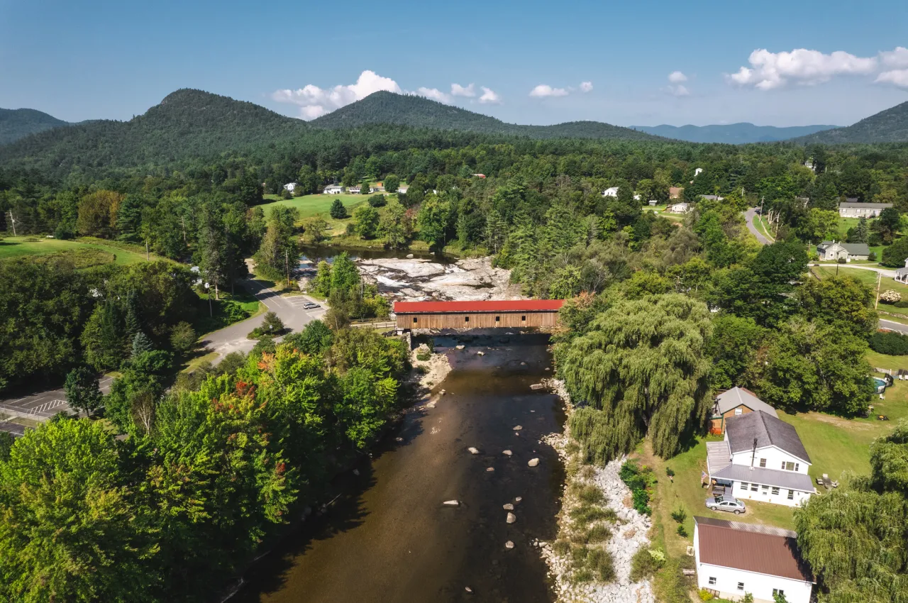 Aerial view of river with covered bridge, houses, and mountains in distance during summer