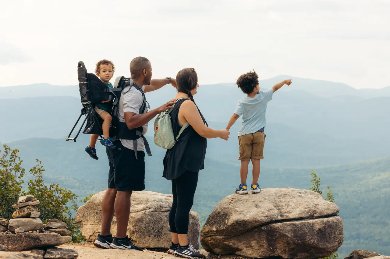 Family on summit, father with baby in backpack and mother holding hand of small child standing on rock