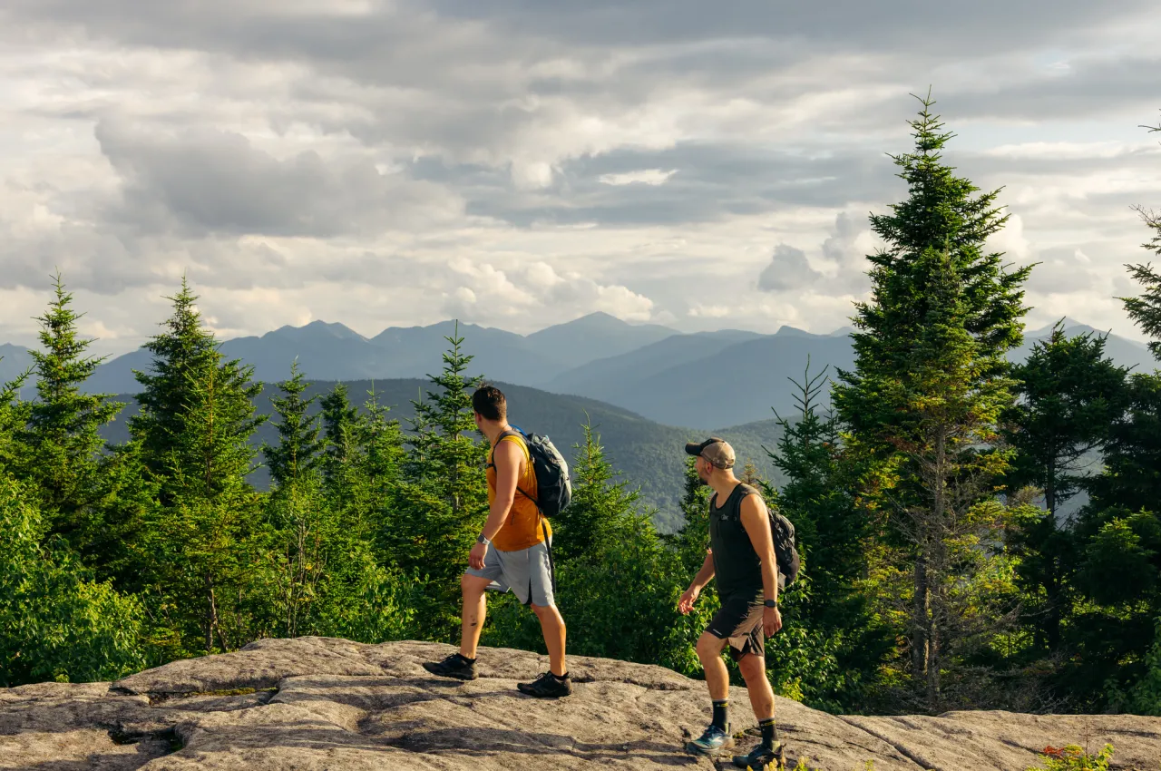 Two men in shorts, tank tops, and hats walk on rocky summit looking at mountain view.