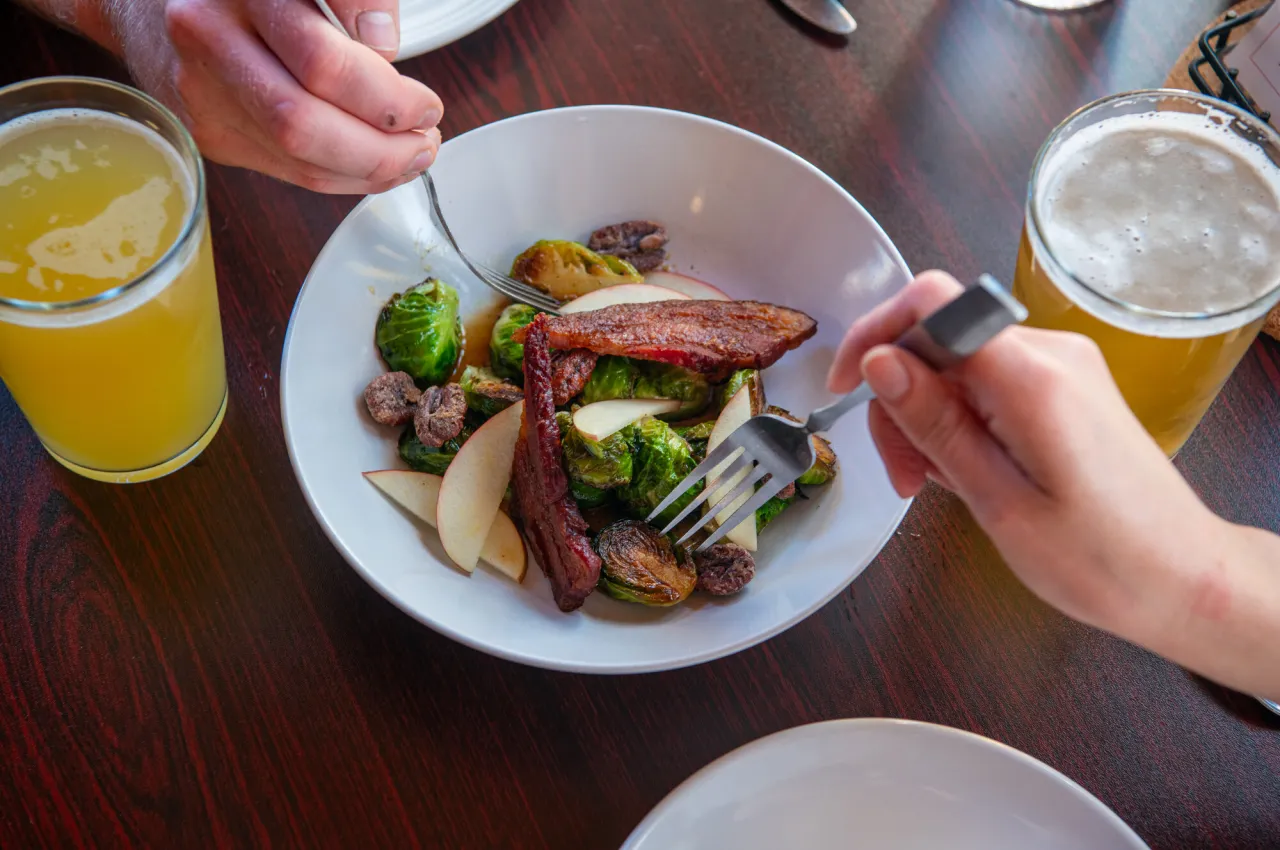 Two people eat a plate of pork and brussel sprouts with drinks
