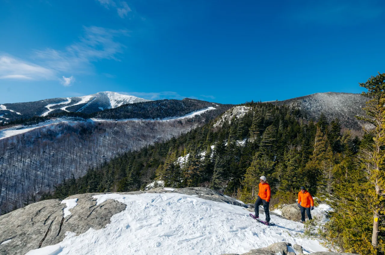 Two men in orange jackets walk up a mountain top in winter.