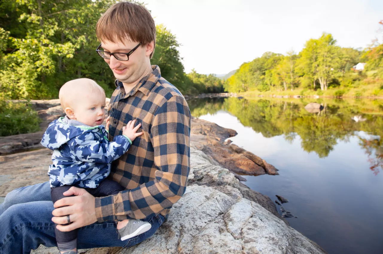 A young man wearing glasses and a plaid shirt holds a cheerful-looking baby while sitting on a rock on the banks of a smooth, tree-lined river.