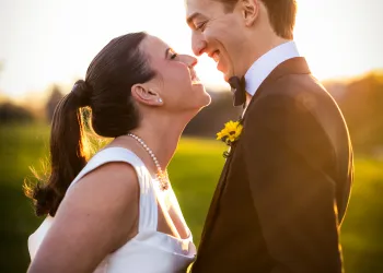 A bride and groom smile at each other in the golden hour of sunlight setting over a lush field