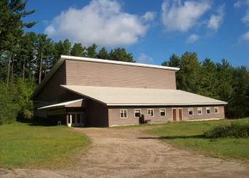Dirt driveway leading to lawn and brown multi-pitched roof building with many windows.
