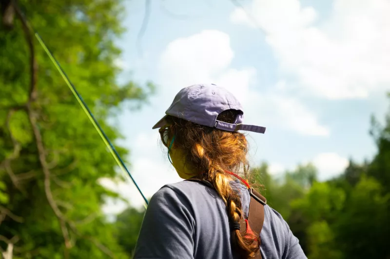 The back of a woman's head; the woman is wearing a baseball cap and is carrying a fly rod.