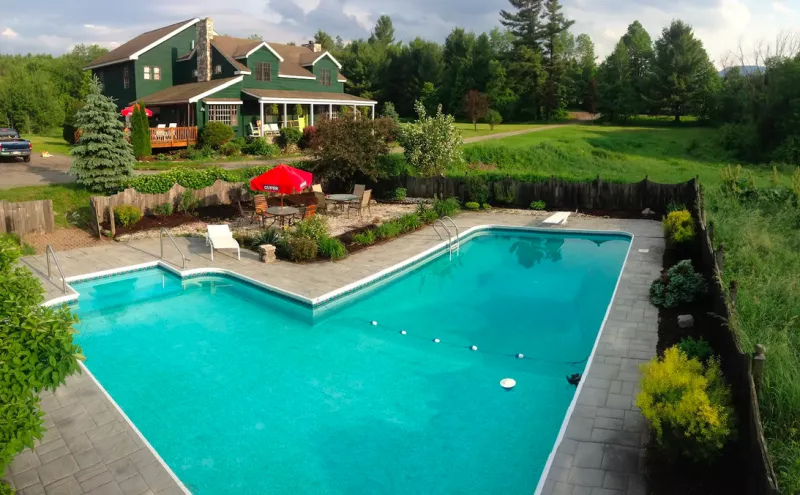 The exterior of a green farmhouse with an in-ground pool and patio in the foreground.