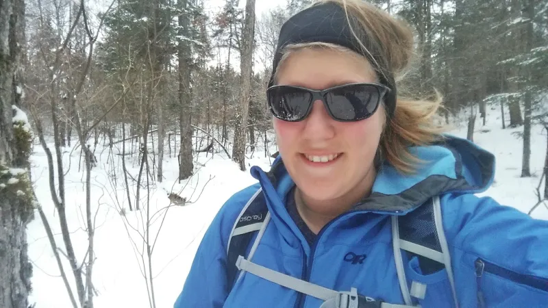 A woman smiling while hiking wearing a blue coat and sunglasses.
