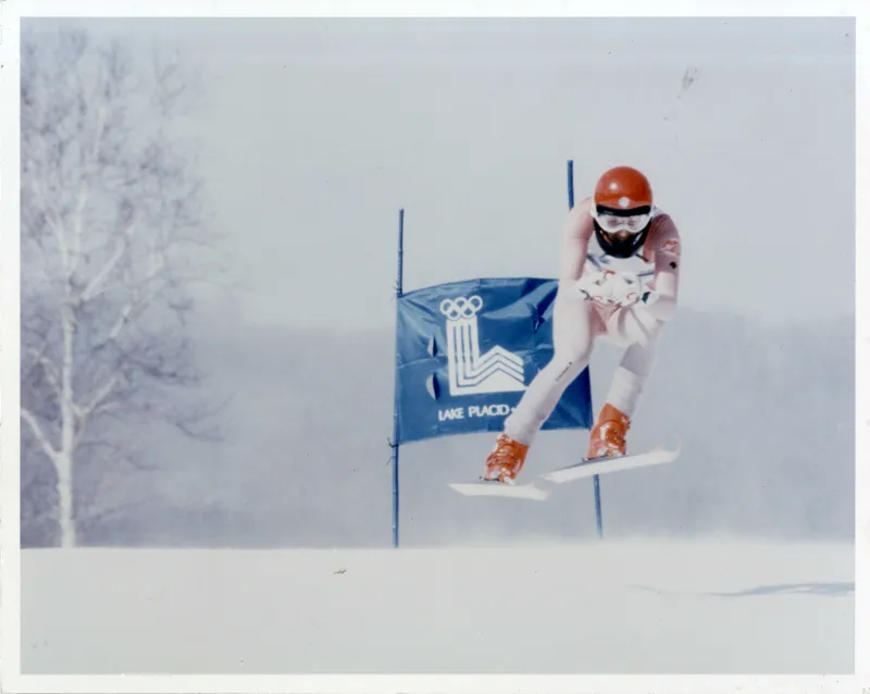 A vintage color photo shows a man in ski competition clothing and helmet competing on a downhill course during the 1980 Olympic Winter Games.