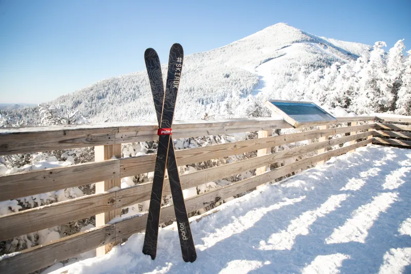 A pair of skis lean against a railing on a deck below snow-covered peaks.