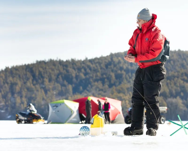 A person in a red jacket stands prepping a line to go in the whole in the ice. Their shanty and friends stand in the background.