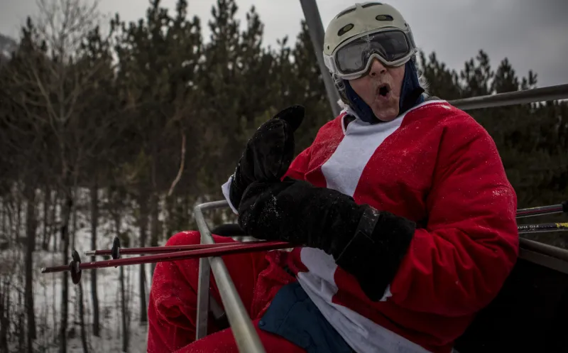 Santa Claus looks surprised on a ski chairlift, in his red suit and ski helmet.