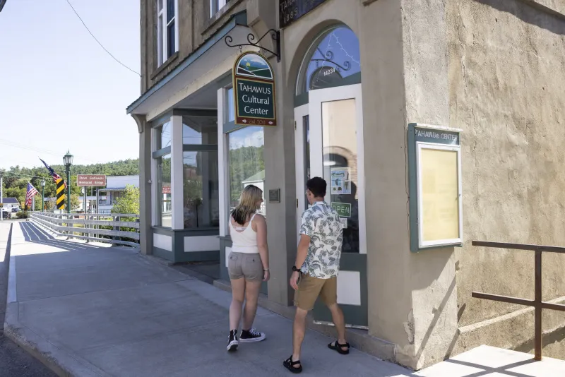 Two visitors enter the Tahawus Center from the main street entrance. Behind them is the sidewalk and a bridge. The Center is beige in color and has a sign that hangs above their door.