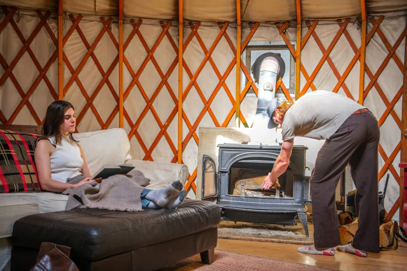 A man fills a wood stove while a woman reads a book in a yurt.
