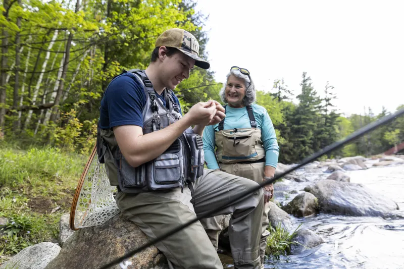 A man and woman smile while the man works on fly fishing gear at a river's edge.