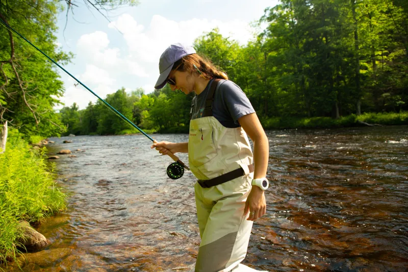 A woman in waders emerges from a river on a sunny day.
