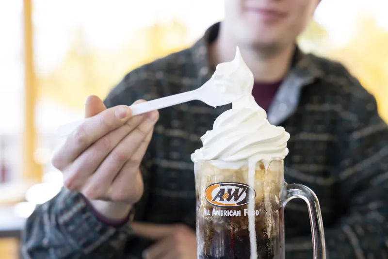 A close up of a root beer float, with a human hand holding a spoon above it.