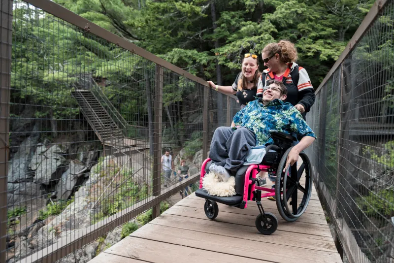 Two women stand next to a man in a wheelchair on a pedestrian bridge; all three are laughing happily.