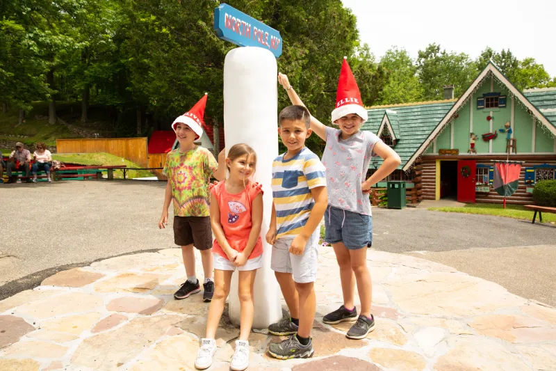 A group of children, some wearing Santa hats, cluster around a pole made of ice on a summer day at a children's theme park.