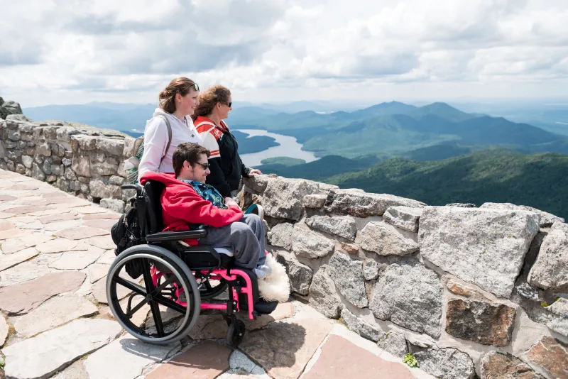 Two women stand next to a young man in a wheelchair; all three are at a scenic viewpoint high on a mountain.