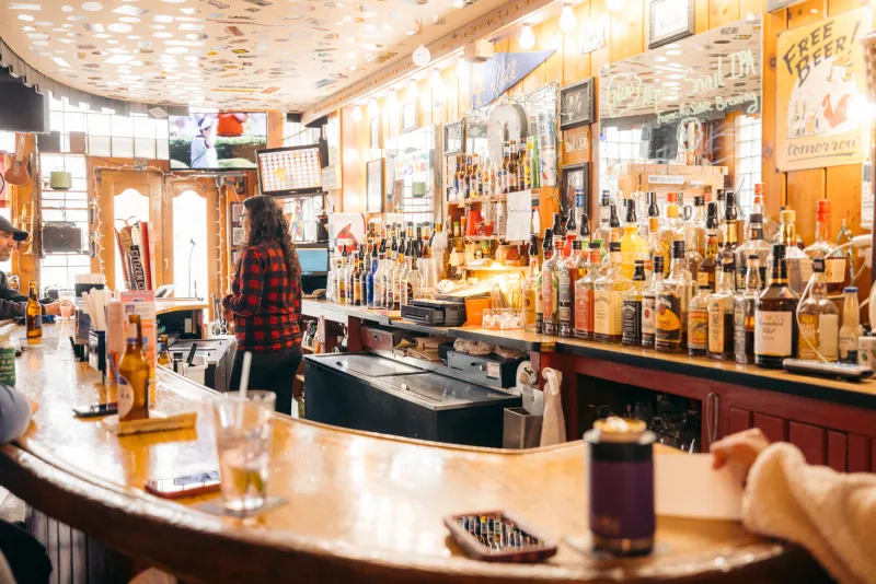 A cheerfully lit, classic bar is decorated with bottles, stickers on the ceiling, and signs filling the walls.